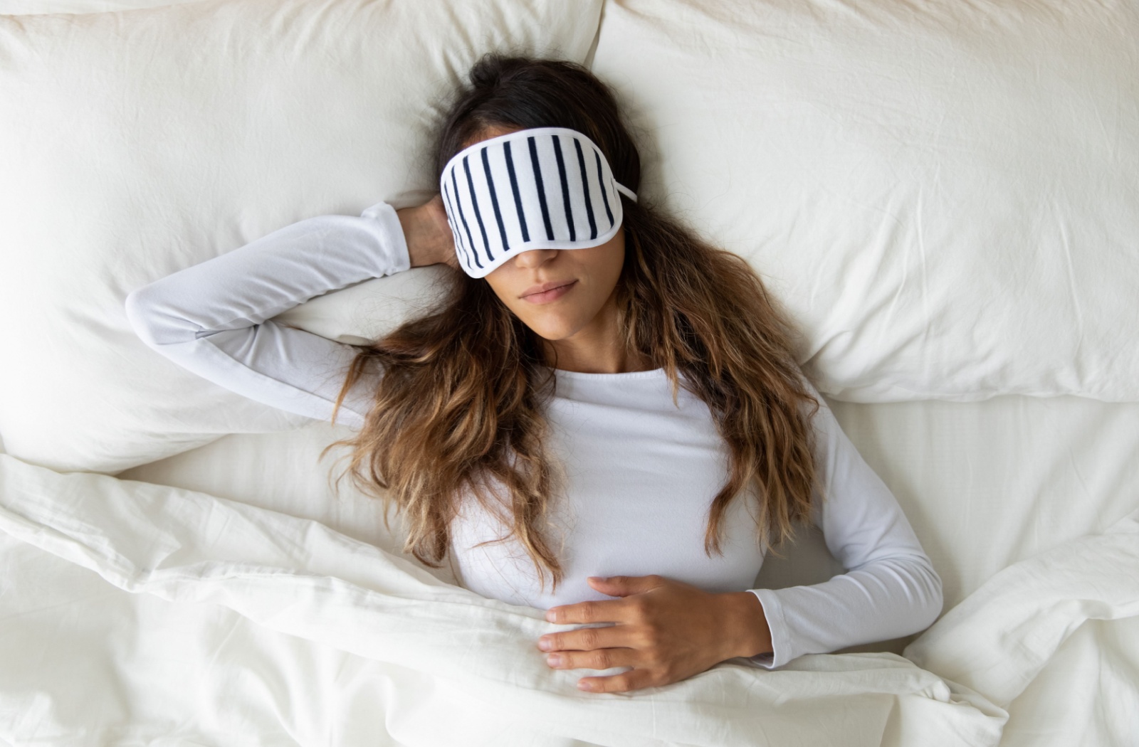 A young adult rests in bed with a striped black and white eye mask on to relieve dry eye symptoms in the middle of the day